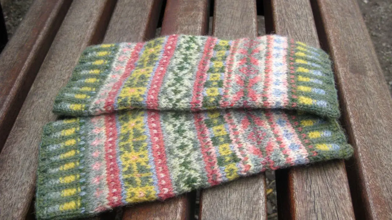 Två handledsvärmare i fair isle stickning med mönster i grönt, rosa, gult och blått ligger på en träbänk.