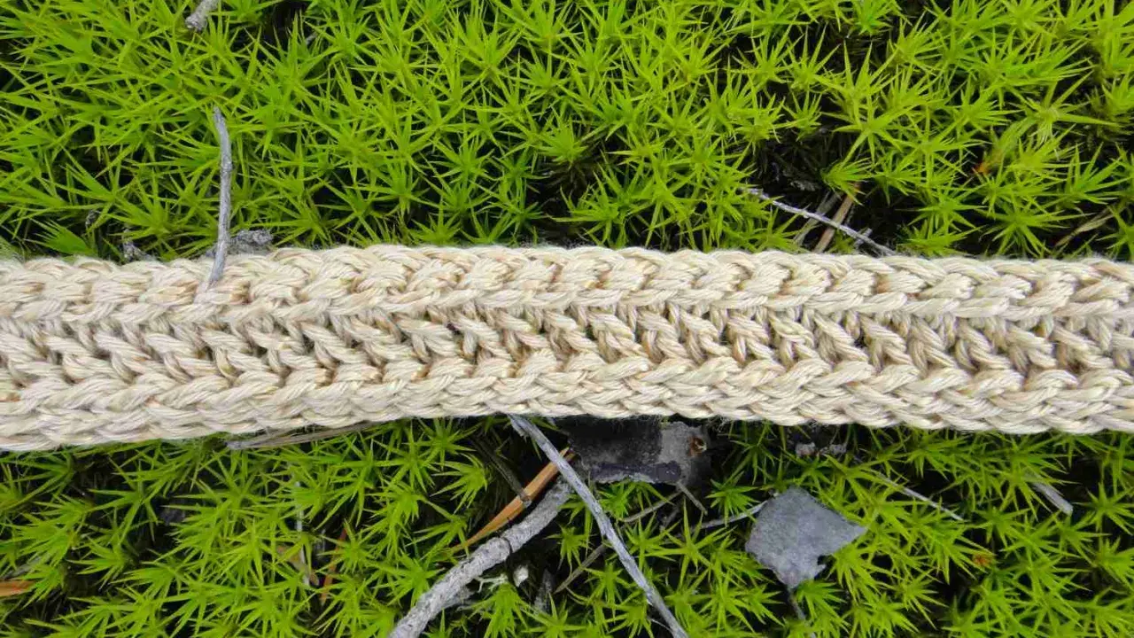 Ett naturfärgat virkat band ligger på grön mossa. Bandet har ett tydligt flätmönster.