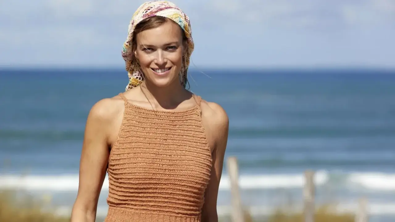 Woman in a crocheted top and headscarf smiles at the camera on a beach.