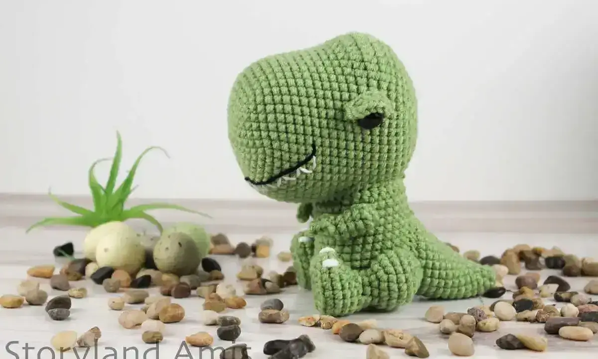 A green crocheted dinosaur sits on a white surface surrounded by small stones and a small plant.