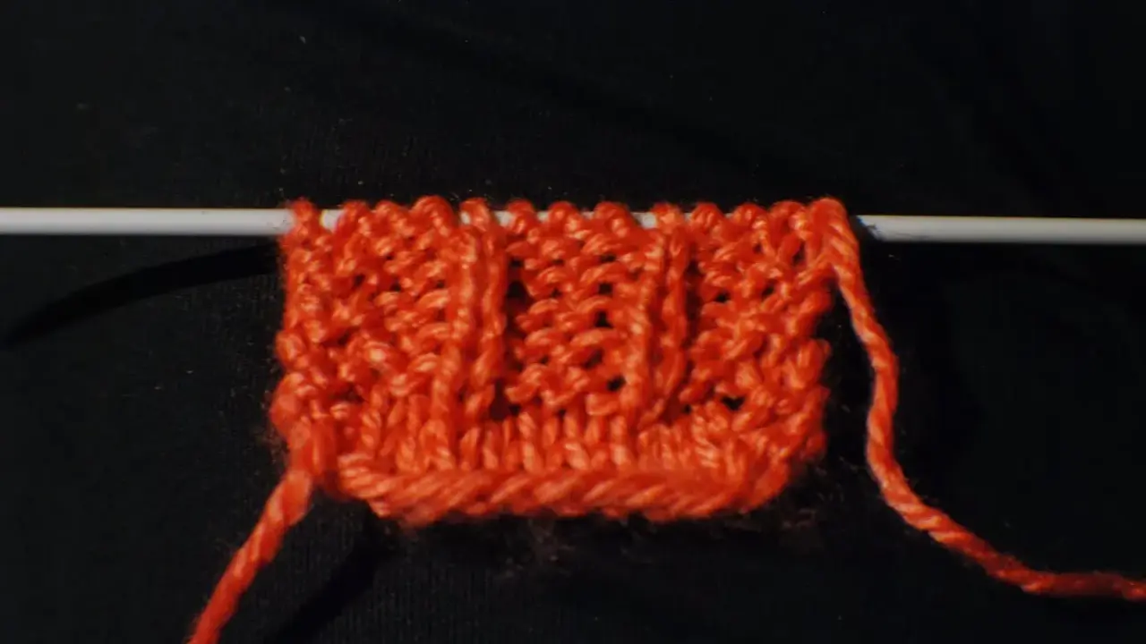 En orange stickad provlapp med ett mönster av vriden resår på en sticka. En lös tråd hänger ner.