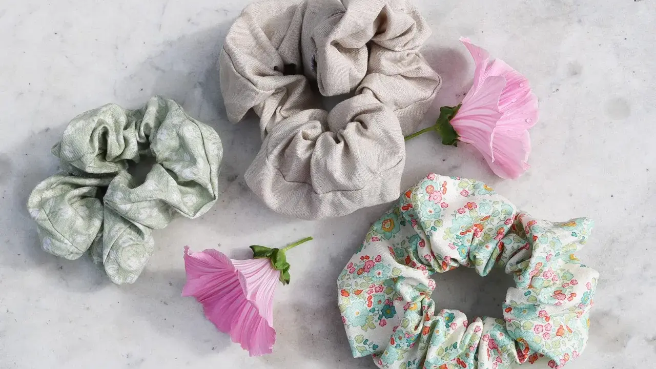 Tre virka scrunchies i olika mönster och färger, omgivna av rosa blommor på en marmorerad yta.