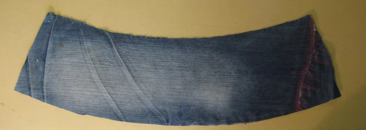 Ett utskuret jeansstycke med en vågig kant, redo att sys till en volang. Röd tråd syns längs ena sidan.