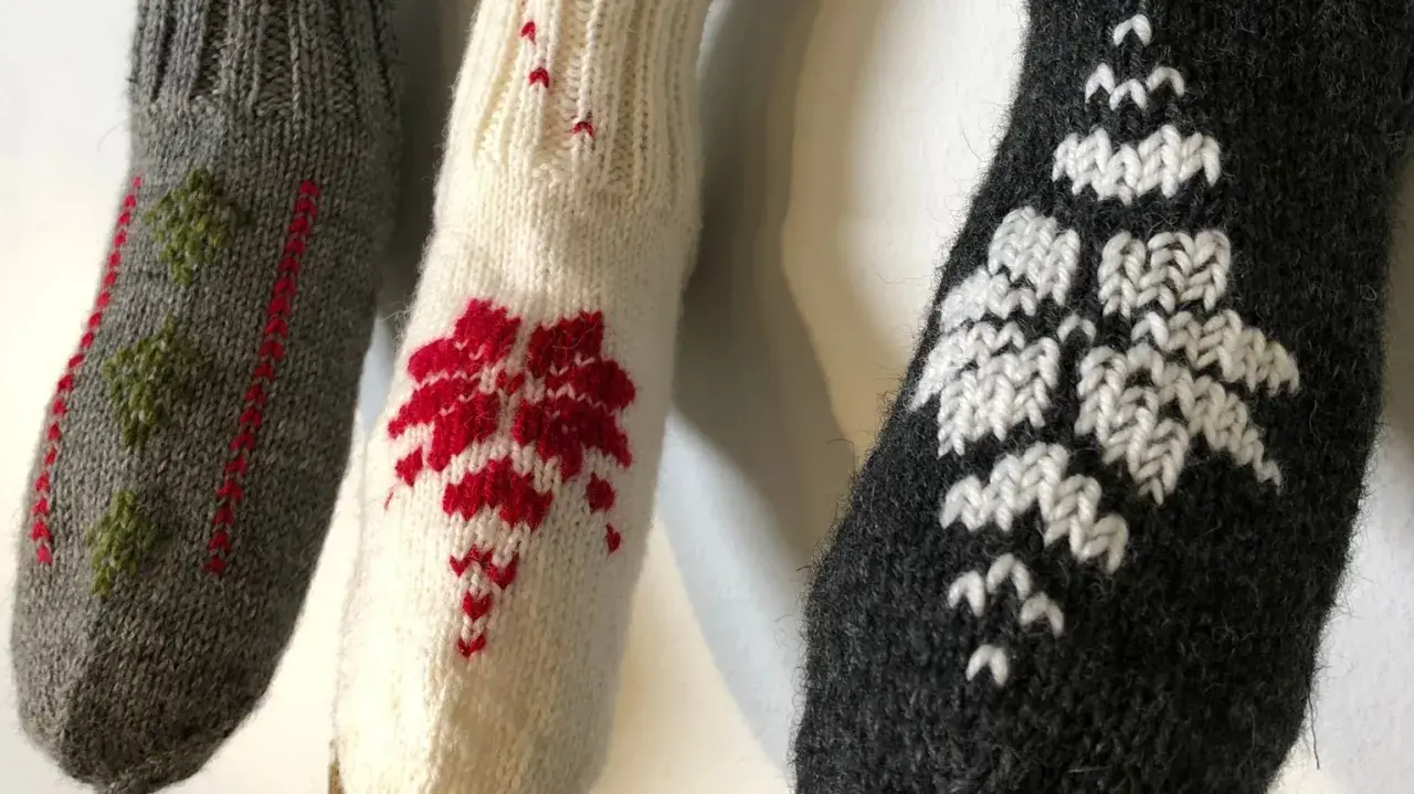 Tre stickade ärmar med mönster i maskstygn. En grå med gröna träd, en vit med röda blommor och en mörkgrå med vita snöflingor.