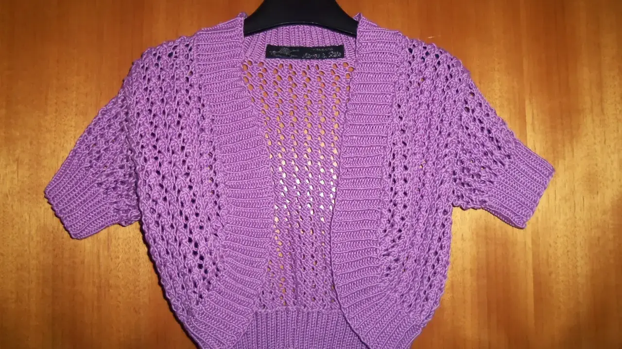 A purple crocheted bolero with short sleeves and an openwork pattern hangs on a hanger against a wooden background.