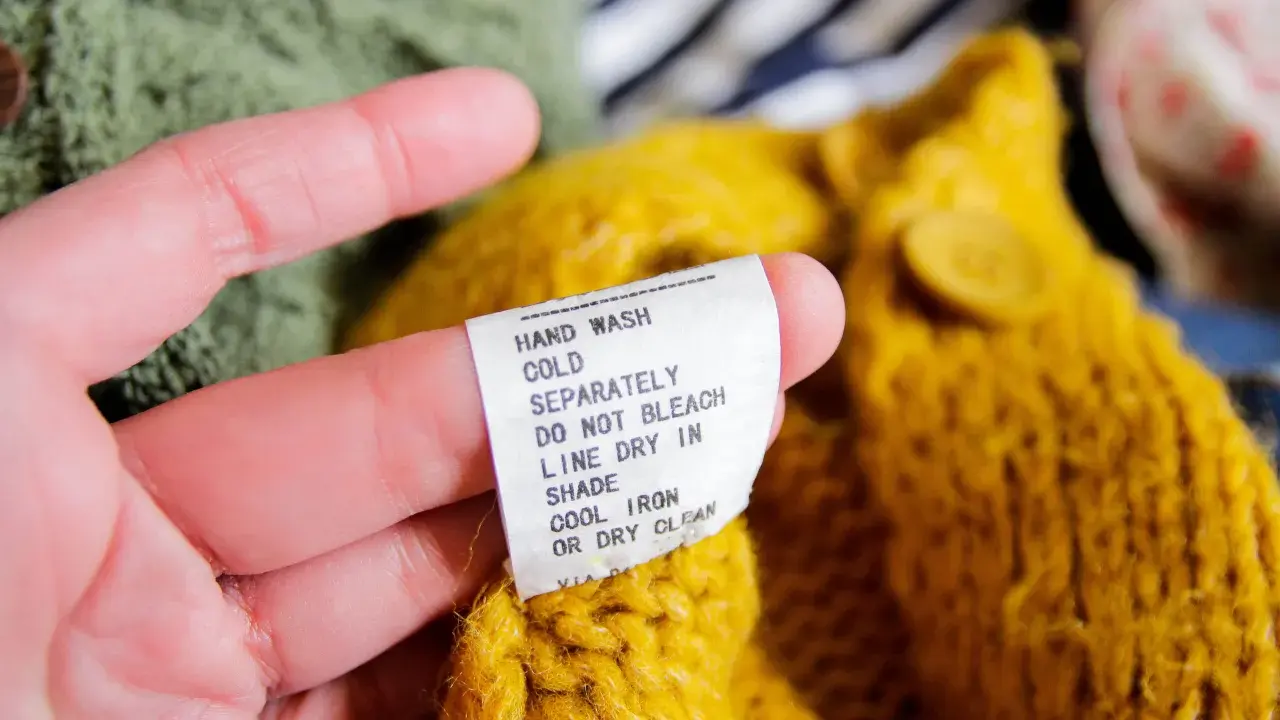 En hand håller i en tvättlapp på en gul stickad tröja. Lappen varnar för att ull kan krympa om den inte sköts rätt.
