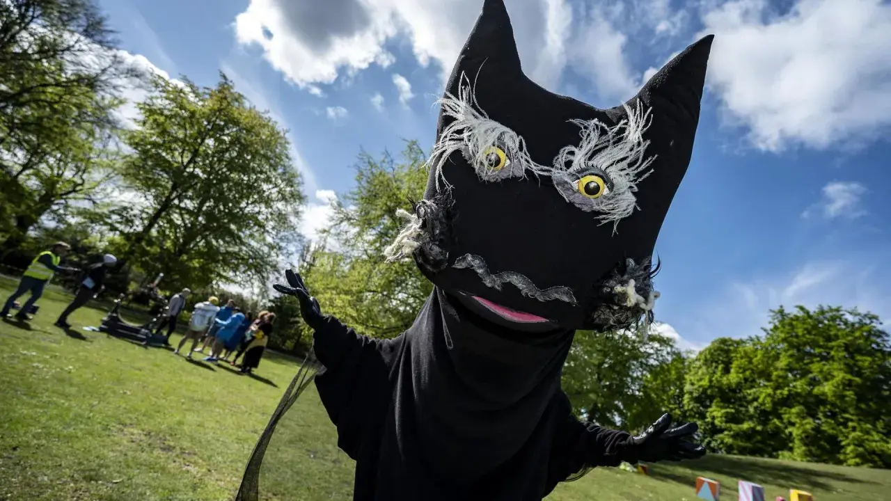 En stor, svart docka som liknar Syrsommarskuggan vinkar glatt i en park med folk i bakgrunden.