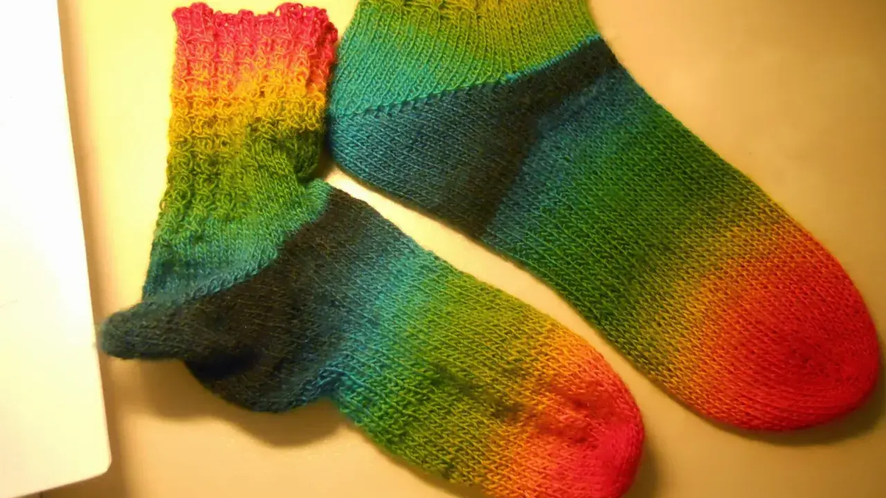 Två regnbågsfärgade, handstickade sockor i ett stycke. En större och en mindre, med färger som går från rosa och gult till grönt och blått.