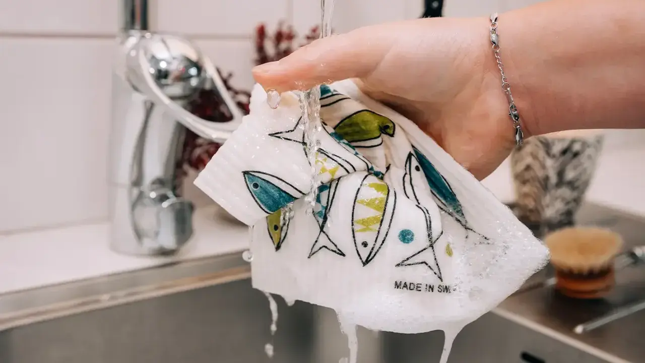 En hand sköljer en disktrasa med fiskmönster under rinnande vatten. Trasan är gjord av garn till disktrasa och har texten "Made in Sweden".
