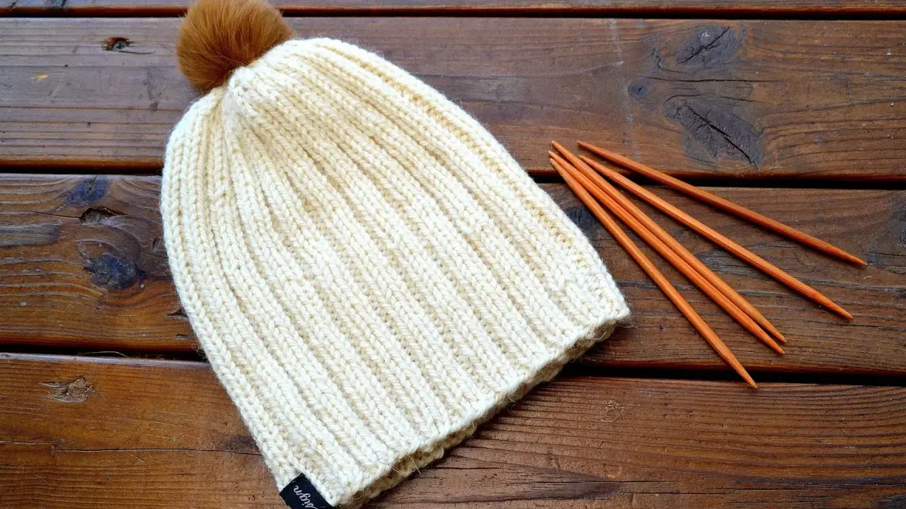 En ribbstickad mössa med en brun pompom ligger på ett träbord bredvid stickor. Perfekt för ett ribbstickad mössa gratis mönster.