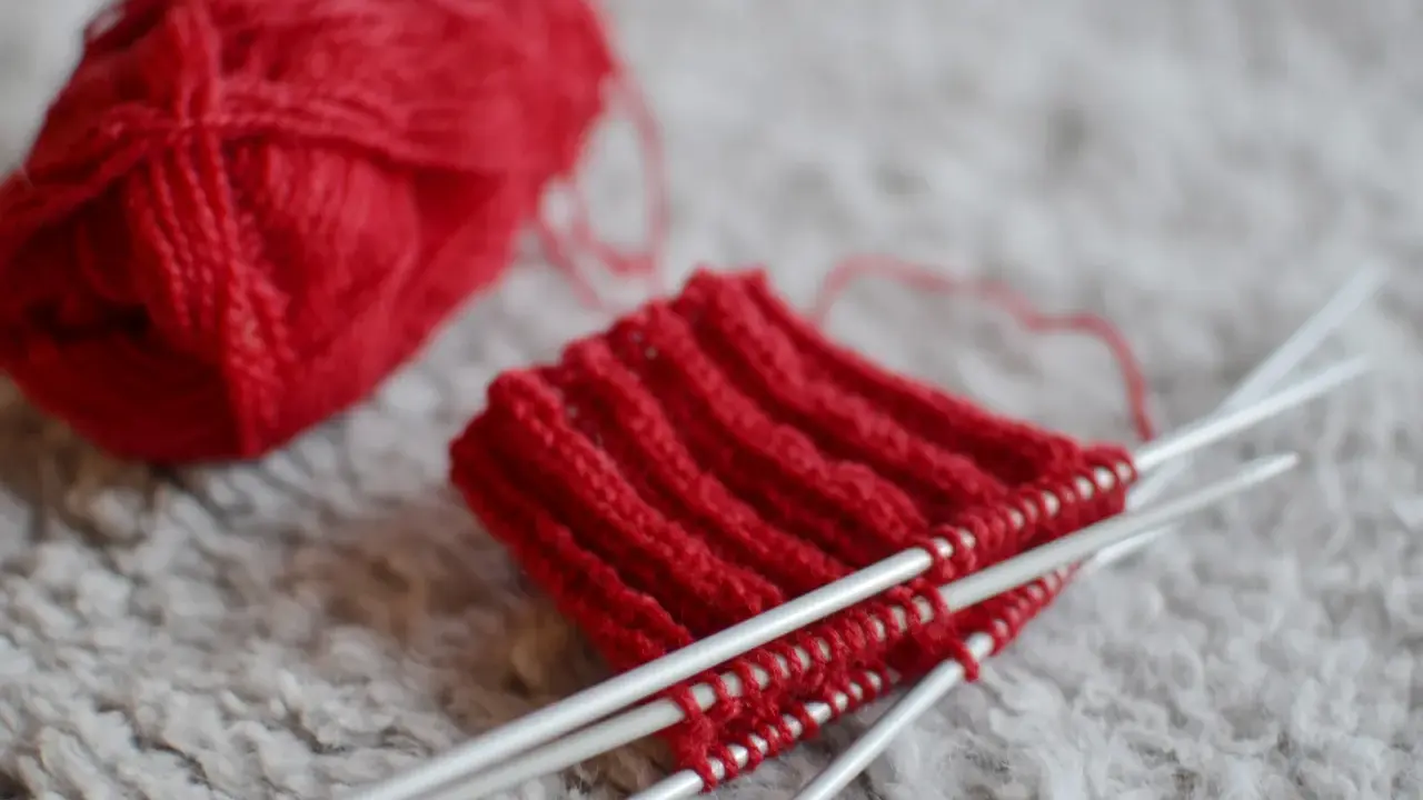 Rött pompom garn ligger bredvid ett stickat provstycke på stickor.
