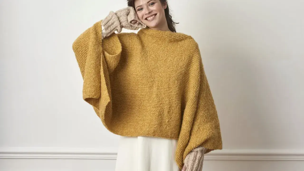 En kvinna ler och håller handen mot ansiktet, klädd i en varm, stickad poncho i senapsgul färg och matchande handledsvärmare.