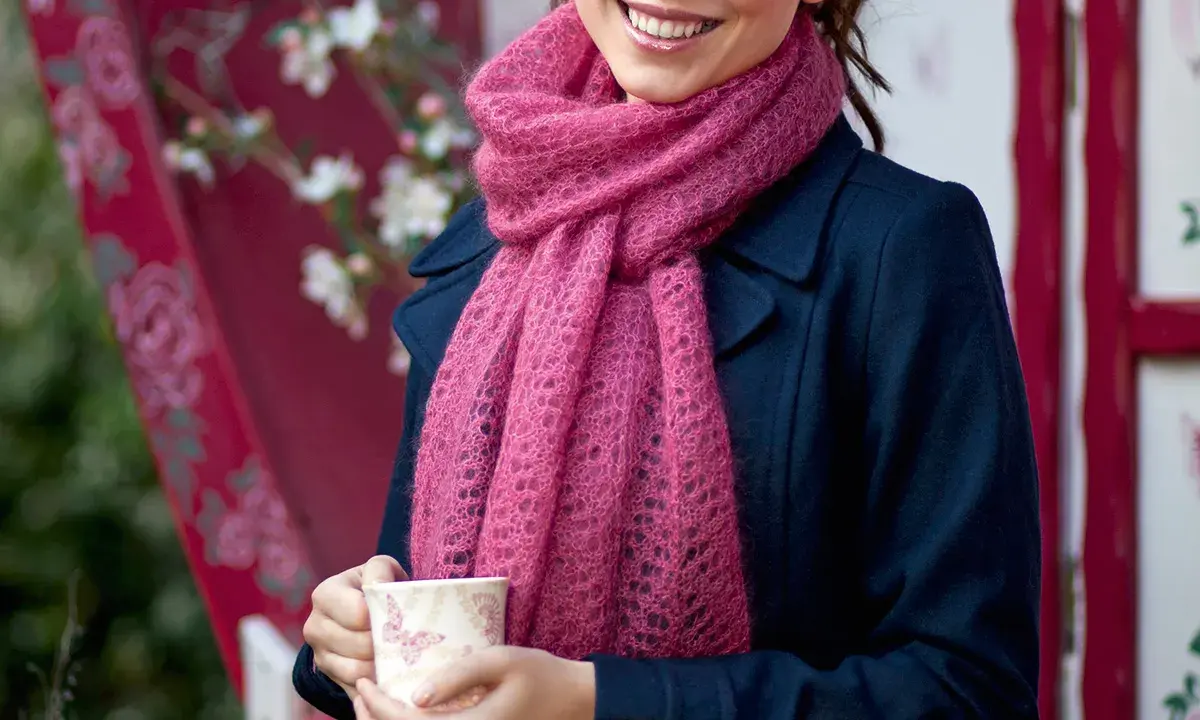 En kvinna ler och håller en mugg, insvept i en mjuk, rosa mohairhalsduk.