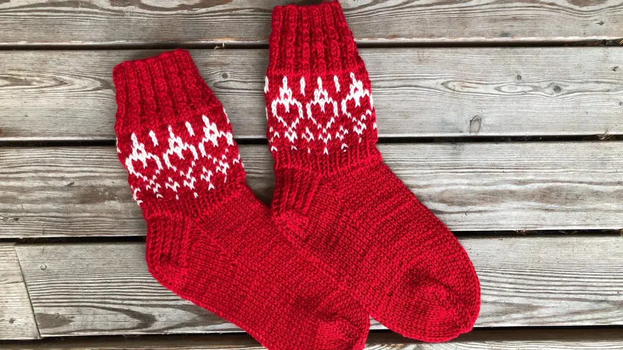 Två par röda, stickade strumpor med vitt mönster av hjärtan och snöflingor ligger på en träbänk. Perfekta för kalla dagar!