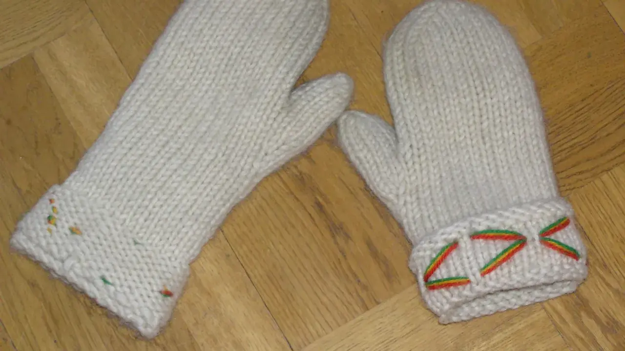 Två vita, stickade vantar med diskreta drops design-mönster i färggranna trådar på muddarna.