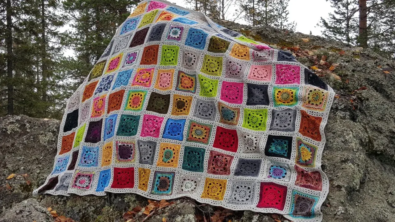 Ett färgglatt, virkat lapptäcke med många olika mönster och färger ligger utspritt över en mossig sten i skogen.