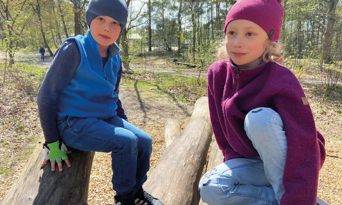 Två barn sitter på stockar i skogen. Båda bär en mjuk merinoull mössa barn, perfekt för utomhuslek.