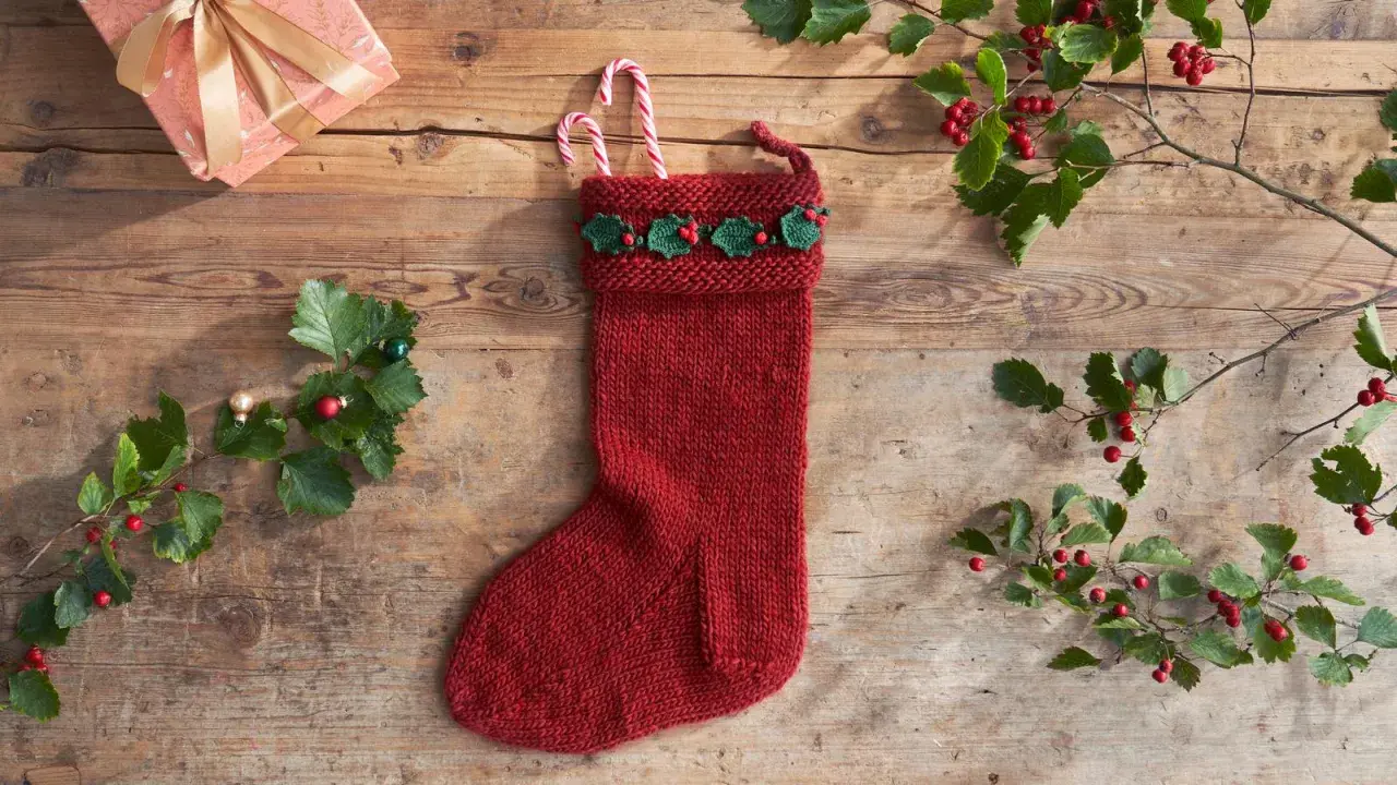 Röd stickad julstrumpa med grön misteldekoration och polkagrisar. En present och julgranspynt ligger på träbordet.