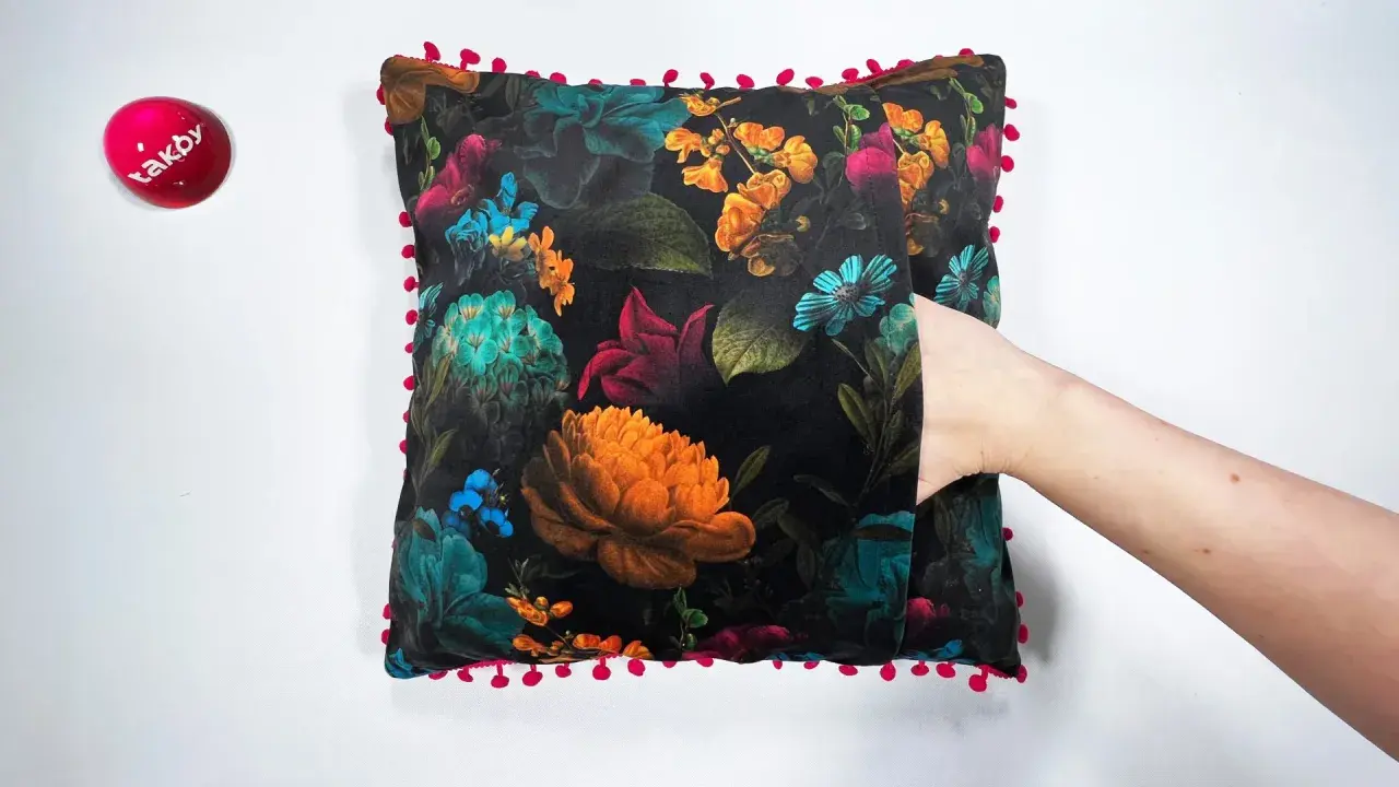 Ett sammetslent kuddfodral med en mörk blommig design och en lekfull kant av små röda bollar. En hand sticker fram ur en ficka på kudden.