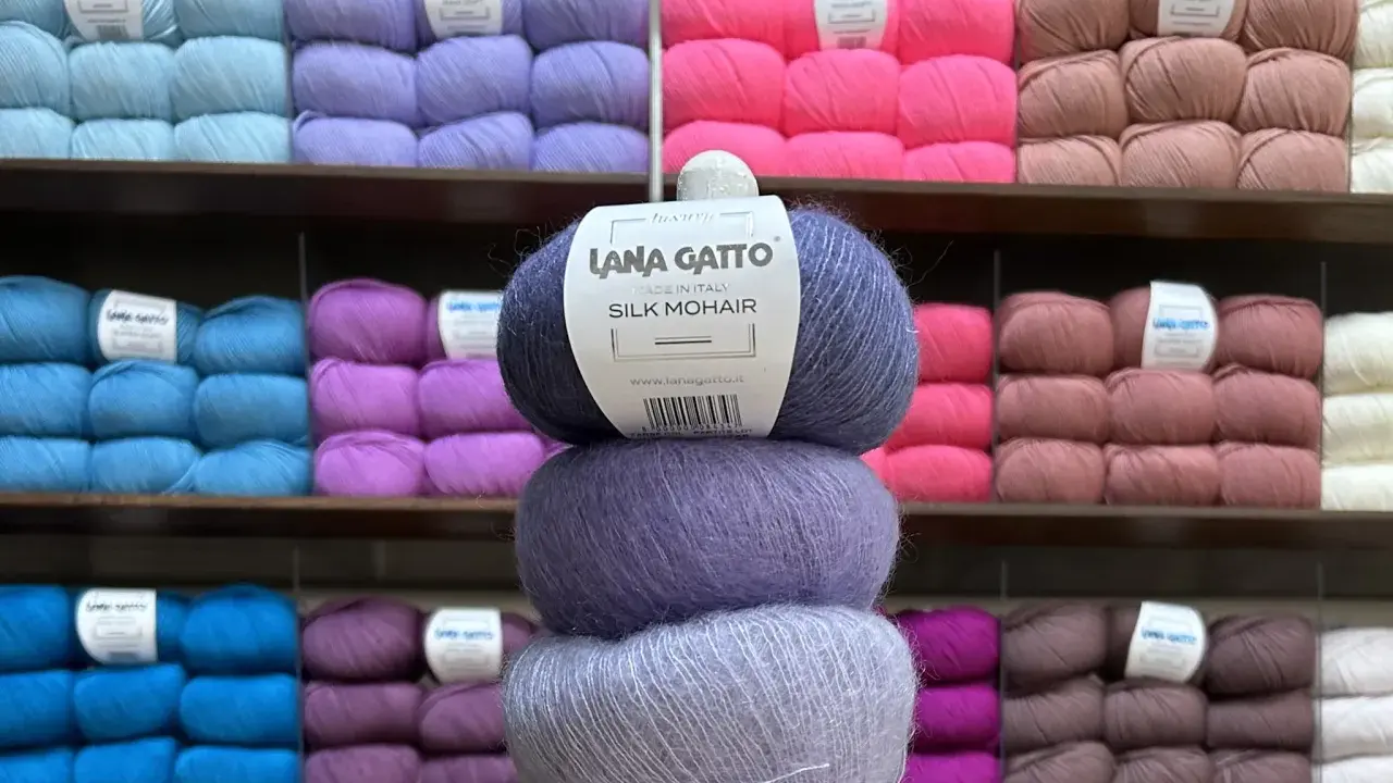 En stapel med tre härvor av lyxigt mohair silke garn i olika lila nyanser. Bakom syns hyllor fyllda med garn i många färger.