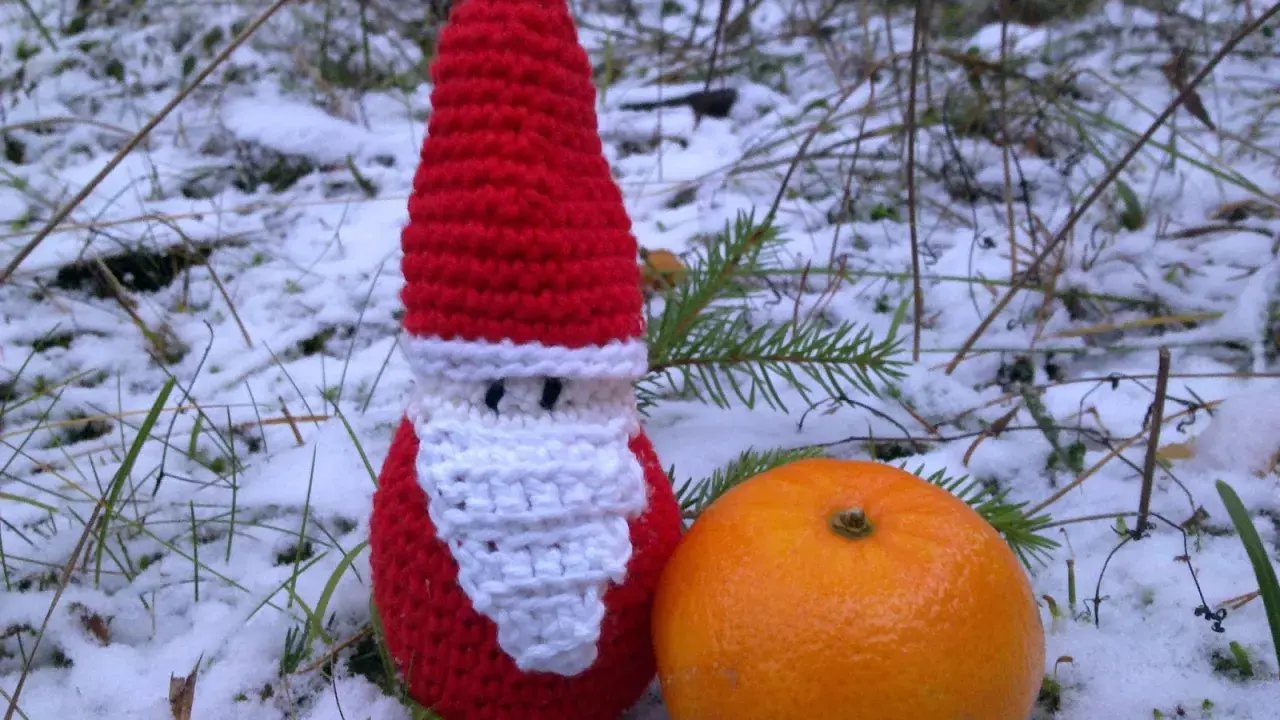 En virkad tomteluva med vitt skägg står i snön bredvid en apelsin.
