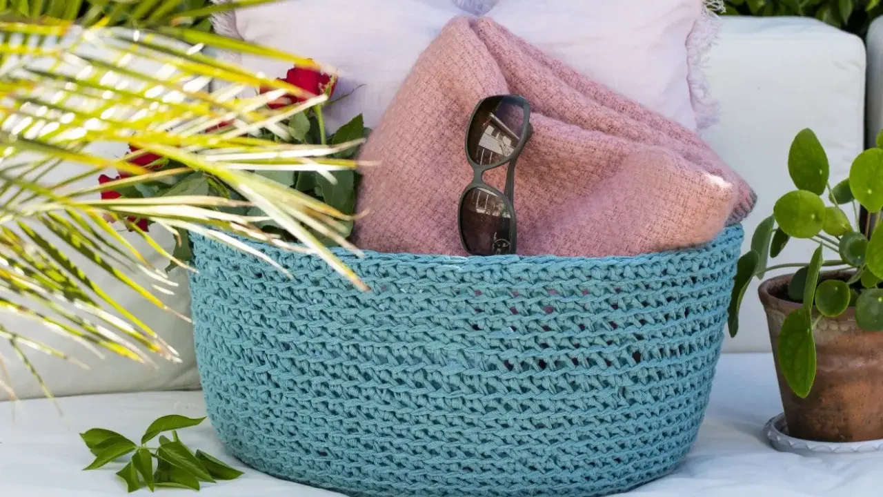 En turkos virkad korg med ett vackert virkmönster, fylld med en rosa filt, solglasögon och röda rosor.