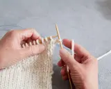 Händer stickar en vit resår med trästickor. En blå kabel syns i bakgrunden.