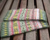 Två handledsvärmare i fair isle stickning med mönster i grönt, rosa, gult och blått ligger på en träbänk.