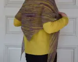 En person visar upp en vackert stickad halsduk i mosstickning med färgskiftningar i gult och lila.