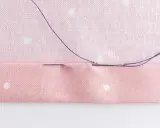 En nål med lila tråd sys genom rosa tyg med prickar. Stygnen är nästan osynliga, ett tecken på skickligt hantverk.