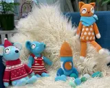 En virkad amigurumi katt med orange och vita prickar sitter på en lurvig matta bredvid en blå mus med tomteluva och en blå elefant i hängselbyxor.