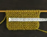 Knitted swatch with a measuring tape across it, showing stitches on wooden needles.