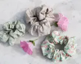 Tre virka scrunchies i olika mönster och färger, omgivna av rosa blommor på en marmorerad yta.