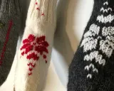 Tre stickade ärmar med mönster i maskstygn. En grå med gröna träd, en vit med röda blommor och en mörkgrå med vita snöflingor.