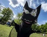 En stor, svart docka som liknar Syrsommarskuggan vinkar glatt i en park med folk i bakgrunden.