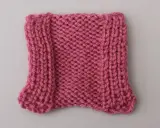 En stickad provlapp i rosa garn, med en vriden maska längs kanterna som ger en vågig effekt.