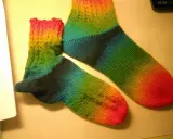 Två regnbågsfärgade, handstickade sockor i ett stycke. En större och en mindre, med färger som går från rosa och gult till grönt och blått.
