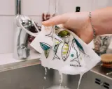 En hand sköljer en disktrasa med fiskmönster under rinnande vatten. Trasan är gjord av garn till disktrasa och har texten "Made in Sweden".