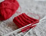 Rött pompom garn ligger bredvid ett stickat provstycke på stickor.