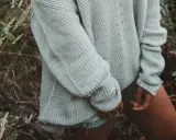 En kvinna i en stickad pullover dam i ljusgrått står i en skog.