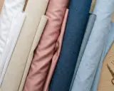 Rullar av tyg i olika färger, inklusive vitt, beige, rosa och blått. Bomullsmaterialet ligger redo för sömnad.