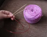 En hand håller i en rundsticka med ett nystan i lila toner bredvid. Dags att sticka resår på rundsticka!