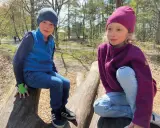 Två barn sitter på stockar i skogen. Båda bär en mjuk merinoull mössa barn, perfekt för utomhuslek.
