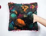 Ett sammetslent kuddfodral med en mörk blommig design och en lekfull kant av små röda bollar. En hand sticker fram ur en ficka på kudden.