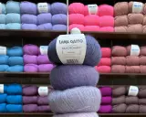 En stapel med tre härvor av lyxigt mohair silke garn i olika lila nyanser. Bakom syns hyllor fyllda med garn i många färger.
