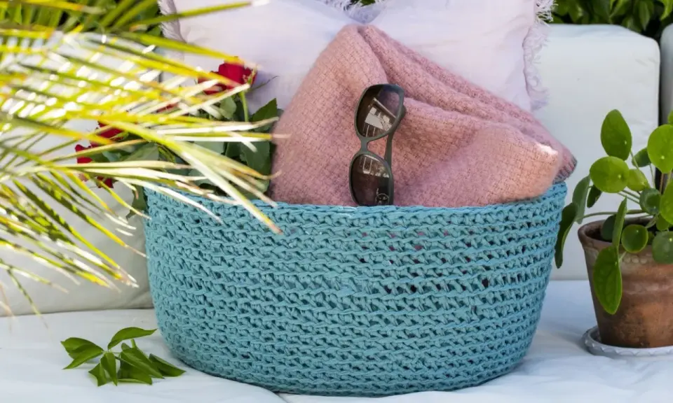 En turkos virkad korg med ett vackert virkmönster, fylld med en rosa filt, solglasögon och röda rosor.