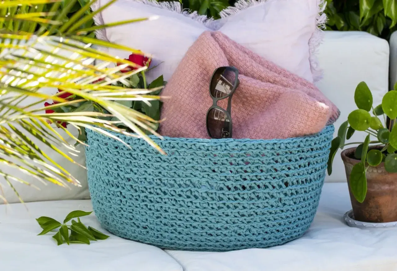 En turkos virkad korg med ett vackert virkmönster, fylld med en rosa filt, solglasögon och röda rosor.