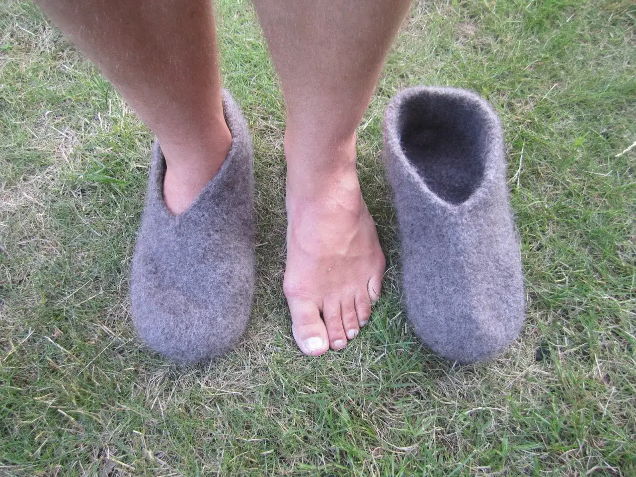 Grå, sticka tovade tofflor står på gräset bredvid ett par bara fötter.