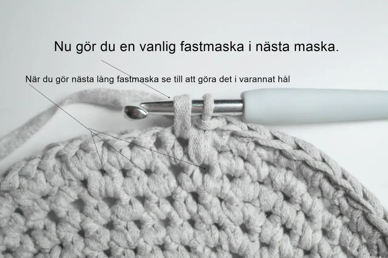 En virkad korg i grått garn med virknål som visar hur man gör en fastmaska.