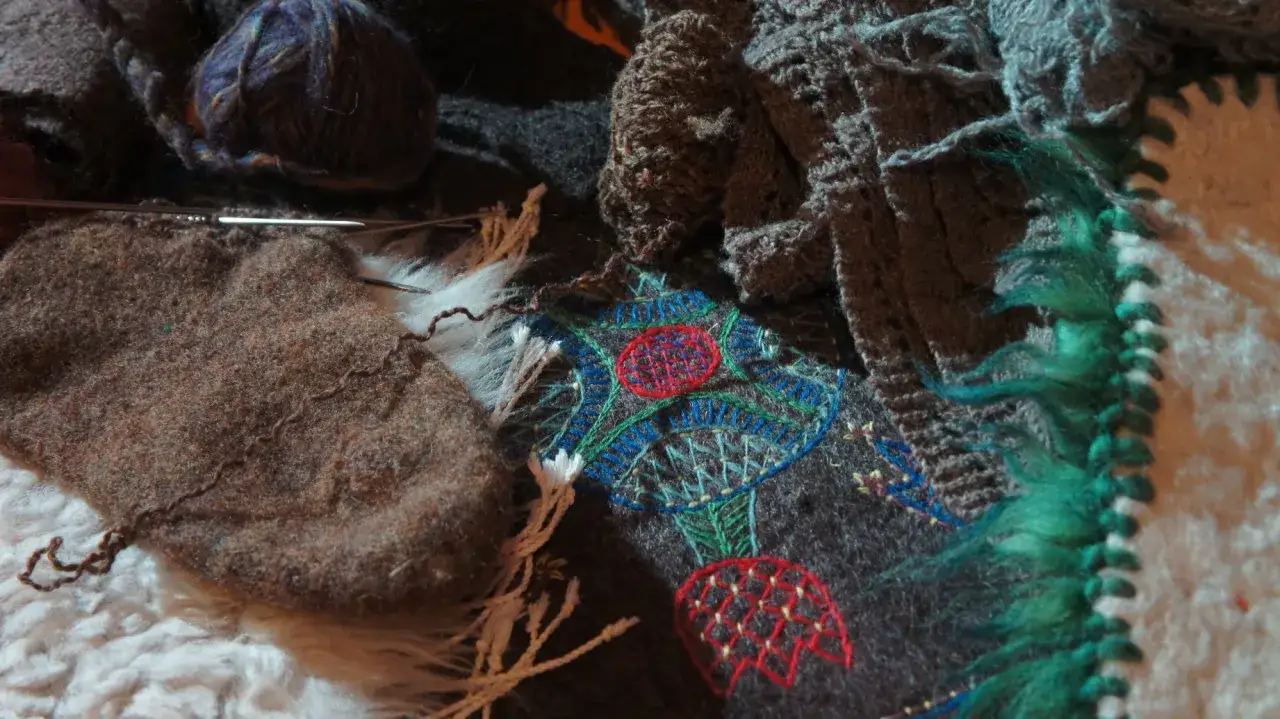 Närbild på karda ull, garnnystan och broderi med blommotiv. Stickor ligger redo för ett nytt projekt.